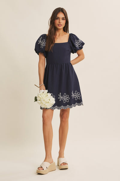A woman modeling a navy blue mini dress with puff sleeves, white floral embroidery, and a scalloped hem, holding a bouquet of white flowers against a solid beige background.