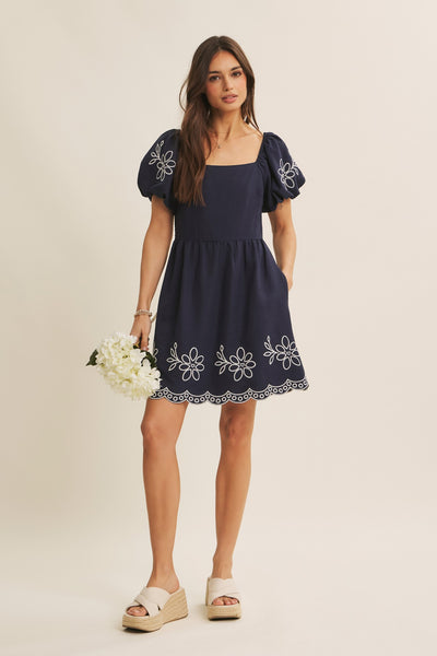 A woman modeling a navy blue mini dress with white floral embroidery on the bodice, sleeves, and hem, paired with beige wedge sandals and holding a bouquet of white flowers against a solid beige background.