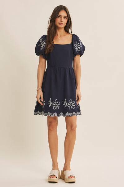A woman modeling a navy blue mini dress with puff sleeves, white floral embroidery, and a scalloped hem, paired with beige platform sandals against a plain beige studio background.