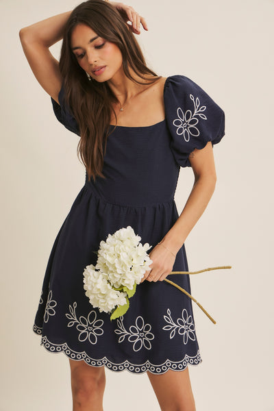 A woman wearing a navy blue puff-sleeve dress with white floral embroidery and a scalloped hem, holding a bouquet of white flowers against a light beige background.