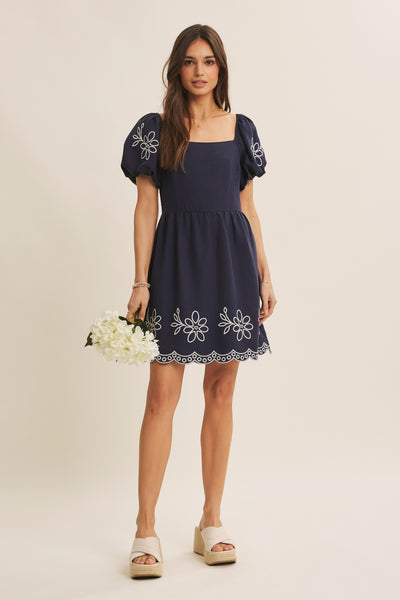 A woman modeling a navy blue mini dress with white floral embroidery on the bodice, sleeves, and hem, paired with cream platform sandals and holding a bouquet of white flowers against a solid light beige background.