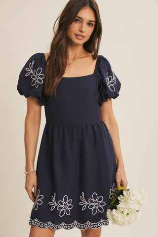 A woman modeling a navy blue mini dress with puff sleeves, white floral embroidery on the sleeves and hem, and a scalloped edge, set against a plain beige background.