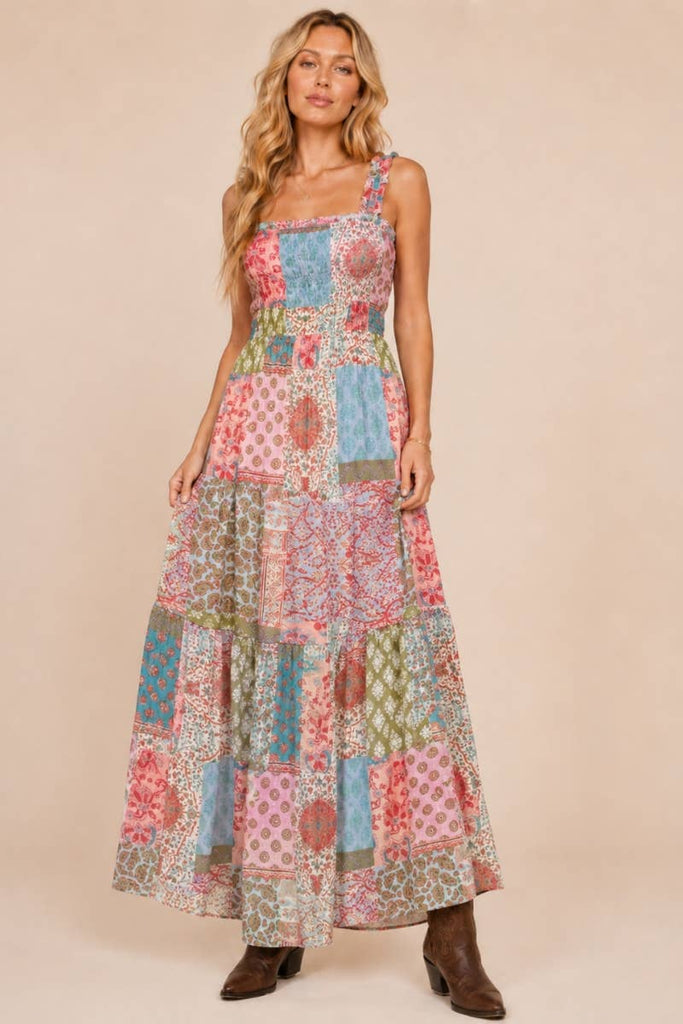 A woman modeling a sleeveless patchwork maxi dress with a multicolor floral and geometric print, paired with brown ankle boots, against a solid beige studio background.
