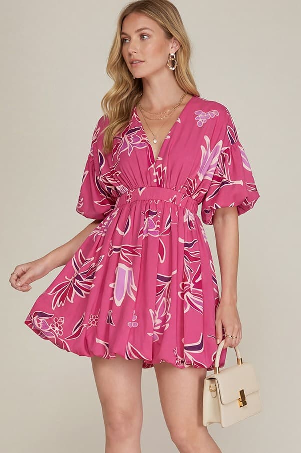 Woman wearing a pink floral dress holding a beige handbag against a plain background