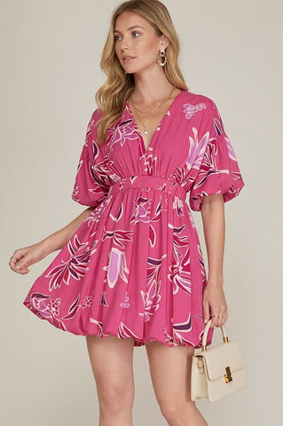 Woman wearing a pink floral dress holding a beige handbag against a plain background