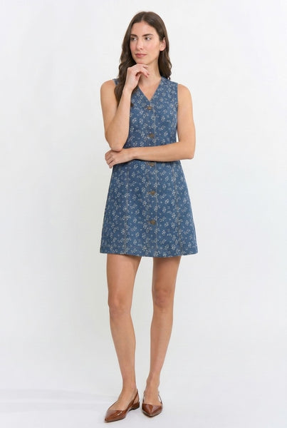 A woman modeling a sleeveless blue mini dress with a small floral print and button-front neckline, standing against a plain light gray studio background.