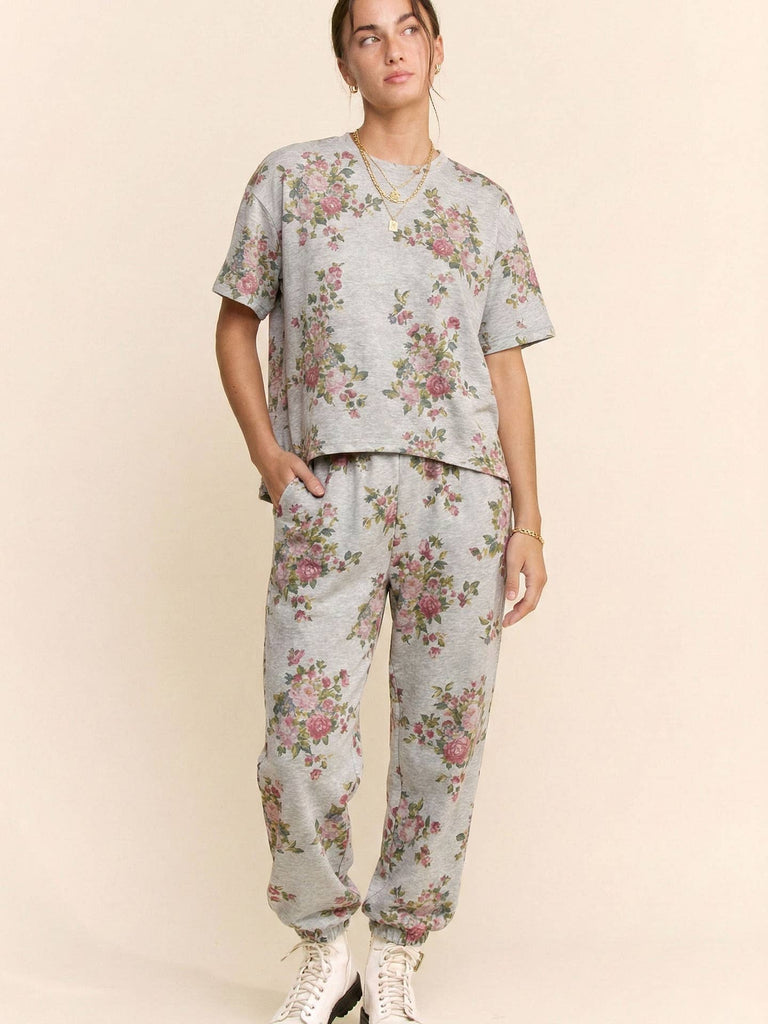 A model wearing a matching light gray floral lounge set, featuring a short-sleeve crewneck top and jogger pants, styled with white sneakers against a solid beige background.