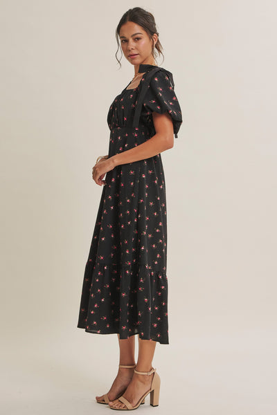 A woman modeling a black midi dress with a small red and pink floral print, featuring puff sleeves and a square neckline with a bow detail, set against a plain light beige studio background.