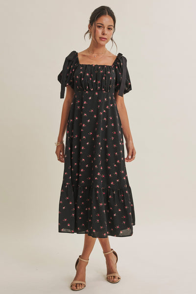A woman modeling a black midi dress with a red and pink cherry print, featuring puff sleeves and a square neckline, set against a plain light beige studio background.