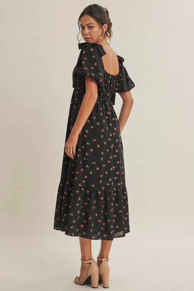 A woman modeling a black midi dress with a small red and pink floral print, featuring ruffled short sleeves and a tie at the back of the neckline, photographed against a neutral light beige studio background.