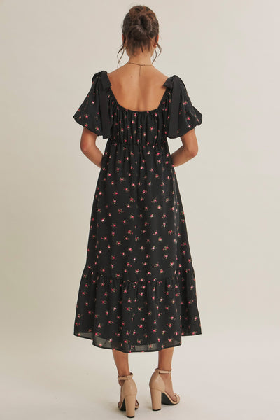 A woman viewed from the back wearing a black midi dress with a small red and pink floral print, puff sleeves, and a square open back, paired with beige high-heeled sandals against a light beige studio background.