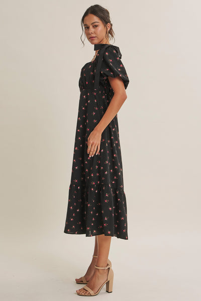 A woman modeling a black midi dress with a small red and yellow floral print, featuring puff sleeves and a high neckline, paired with beige high-heeled sandals against a plain light beige studio background.
