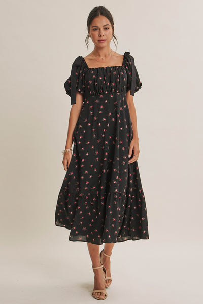 A woman modeling a black midi dress with a small red and pink floral print, featuring puff sleeves and a square neckline, set against a plain light beige studio background.