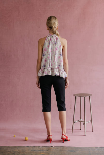 A woman stands with her back to the camera wearing a sleeveless floral halter top, black capri pants, and red heels, posed in front of a textured pink wall with a stool nearby.