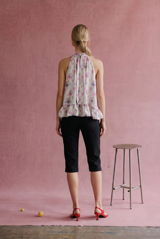 A woman stands with her back to the camera wearing a sleeveless floral halter top, black capri pants, and red heels, posed in front of a textured pink wall with a stool nearby.