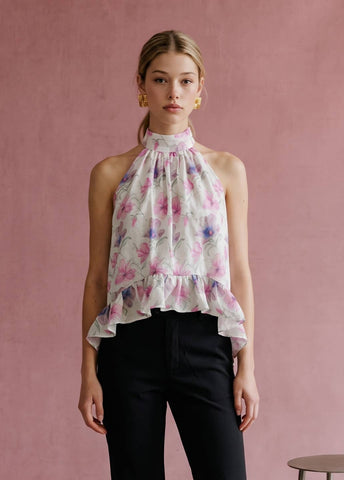 A woman wearing a sleeveless halter-neck blouse with a pink and purple floral print on a white base, paired with black pants, standing against a textured dusty pink wall.