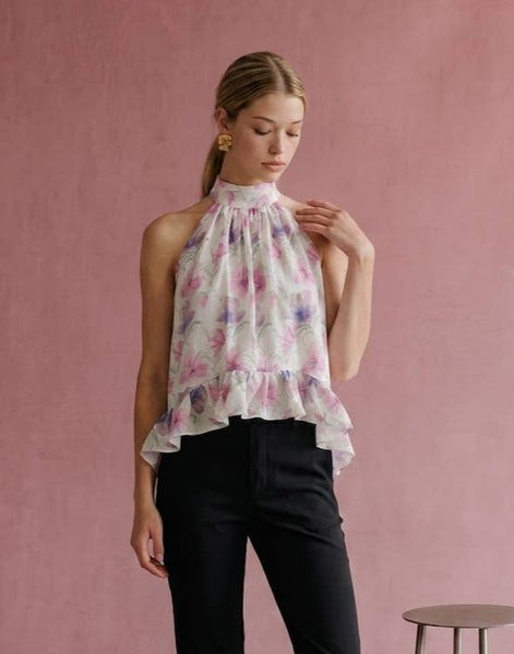 A woman wearing a sleeveless high-neck blouse with a pink and purple floral print on a white base, paired with black pants, standing against a textured pink wall.