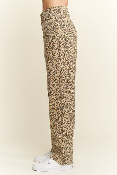 A side view of a person wearing leopard-print trousers and white sneakers against a plain beige studio background.