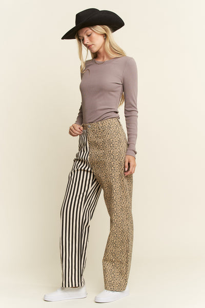 A woman modeling a pair of mixed-pattern wide-leg pants with one leg in black and white stripes and the other leg in a beige leopard print, paired with a mauve long-sleeve top, a black hat, and white sneakers against a solid cream background.