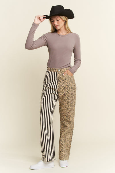 A woman modeling a long-sleeve taupe top, mixed-print pants with black and white vertical stripes on one leg and a tan leopard print on the other, a black cowboy hat, and white sneakers against a solid light beige background.