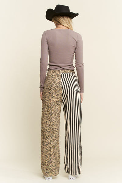 A woman shown from the back wearing a black cowboy hat, a taupe long-sleeve top, and wide-leg pants with a split design: leopard print on one leg and black-and-white vertical stripes on the other. She stands against a plain light beige studio background.