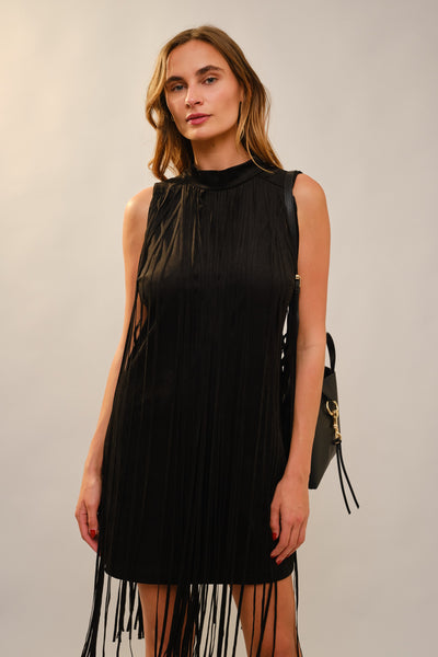 A woman modeling a sleeveless black pleated mini dress with fringe detailing at the hem and shoulders, paired with a black shoulder bag, against a plain light gray studio background.