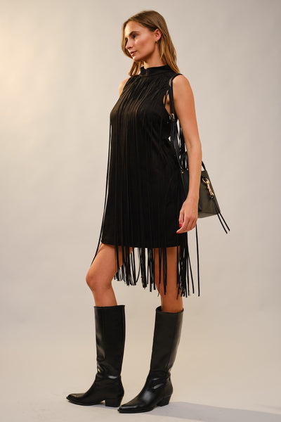 A woman modeling a sleeveless black fringe dress with a high neck, paired with black knee-high boots and carrying a small black handbag, against a plain light gray studio background.