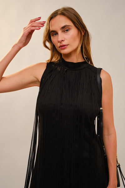 A woman wearing a sleeveless black pleated dress with fringe detailing and a high mock neck, posed against a plain light gray studio background.