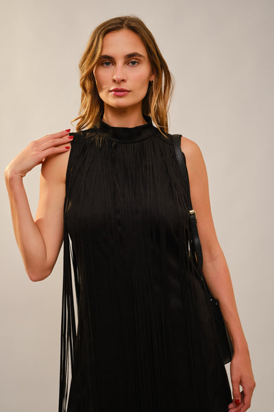 A woman modeling a sleeveless black pleated dress with a high mock neck and long fringe details, photographed against a neutral light gray studio background.
