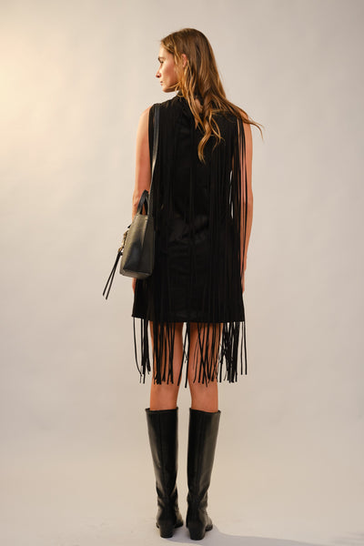 A woman shown from the back wearing a sleeveless black fringed dress with long fringe hanging from the hem, paired with black knee-high boots and a black shoulder bag, against a plain light gray studio background.