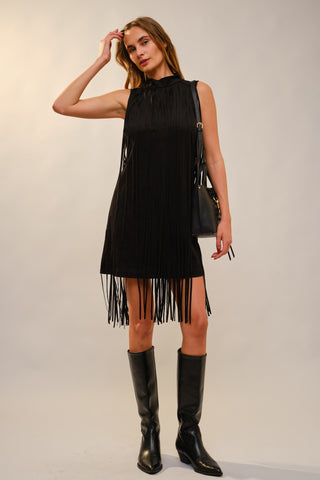 A woman modeling a sleeveless black fringe mini dress with a high neck, paired with black knee-high boots and a black shoulder bag, against a plain light gray studio background.