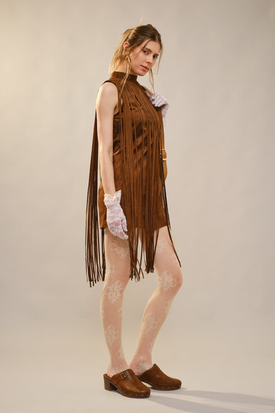A model wearing a brown suede fringe dress with white lace gloves and brown clogs, standing against a neutral beige studio background.