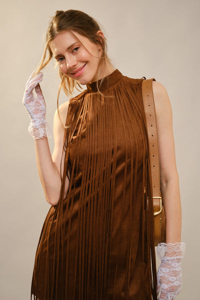 A woman wearing a sleeveless brown pleated dress with fringe, white lace gloves, and a tan shoulder bag, posed against a plain light beige studio background.