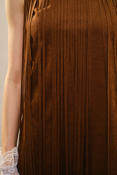 Close-up of a brown pleated sleeveless dress with long fringe, worn by a model whose arm and hand with a white lace glove are visible on the left.