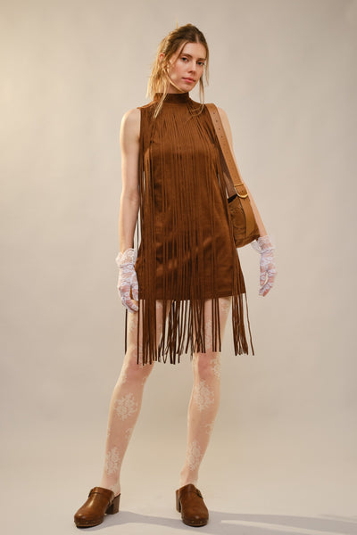 A model stands against a plain beige studio background wearing a sleeveless brown fringe dress with a high neck, sheer white lace gloves, patterned tights, brown clogs, and a brown shoulder bag.
