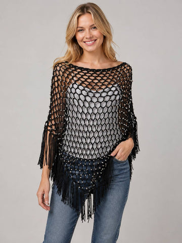 A woman modeling a black open-knit poncho-style top with fringe and bead details over a gray top and blue jeans, photographed against a plain light gray studio background.