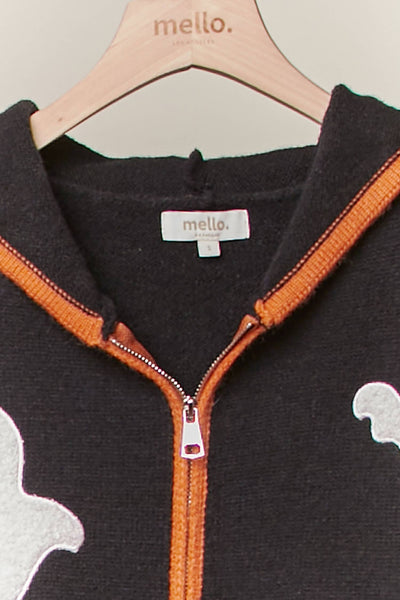A close-up of a black knit zip-up hoodie with orange trim and white ghost graphics, hanging on a wooden hanger branded 'mello.'.