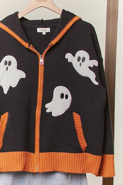 A black knit zip-up hoodie with orange ribbed trim and white ghost motifs, hanging on a wooden hanger against a light wall.