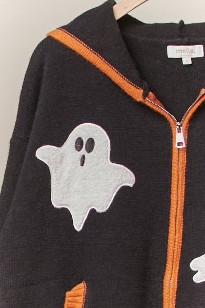 A close-up of a dark charcoal gray knit zip-up hoodie with orange trim and a white ghost applique on the front. The brand label 'mella' is visible inside the collar.