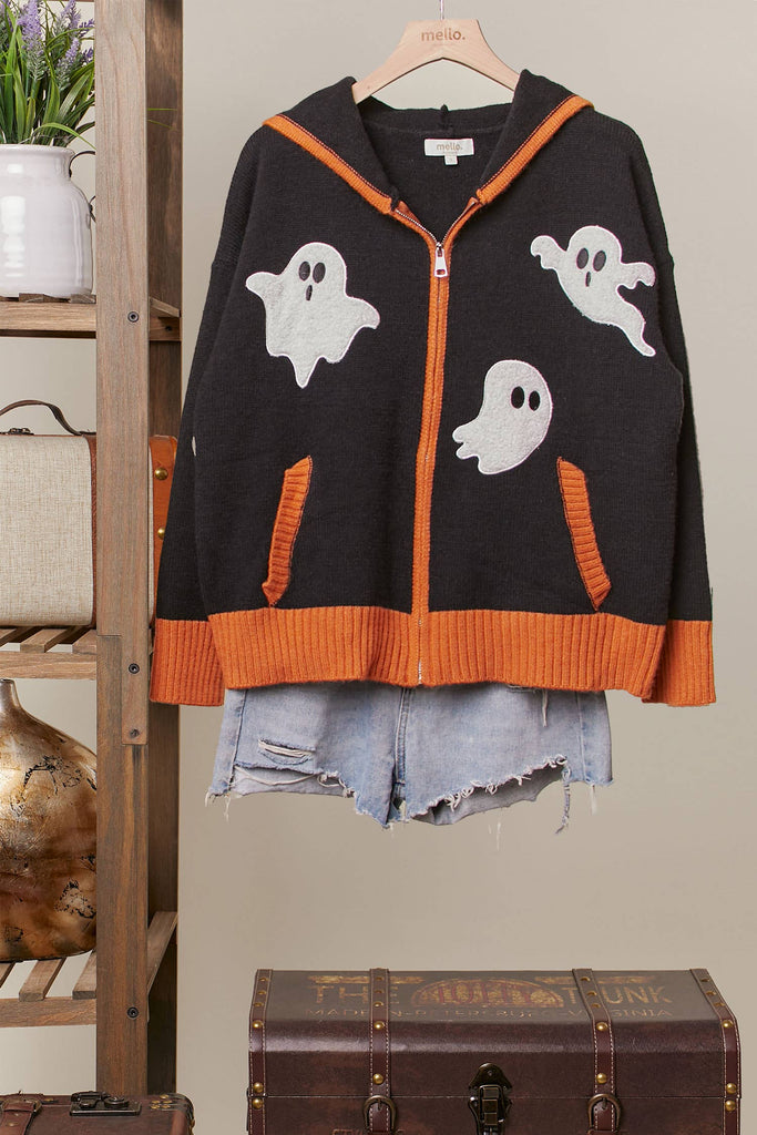 A black knit zip-up hoodie with orange ribbed trim and three white ghost graphics, styled over light blue denim shorts on a wooden hanger against a neutral indoor backdrop.