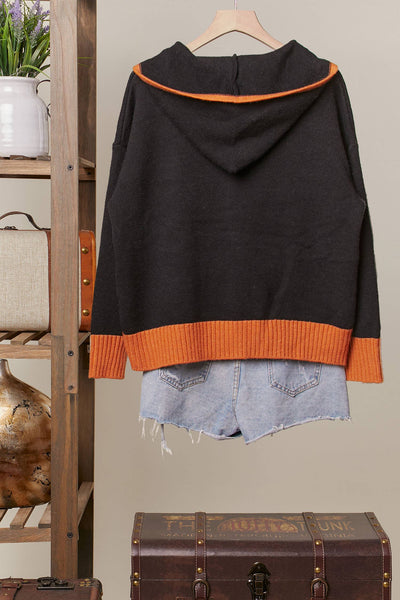 A black hooded sweater with bright orange ribbed trim at the hood and hem, hanging on a wooden hanger. The sweater is styled with light-wash denim shorts underneath, set against a neutral indoor background with a wooden shelf and vintage trunk.
