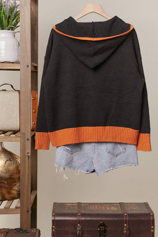 A black hooded sweater with bright orange ribbed trim at the hood and hem, hanging on a wooden hanger. The sweater is styled with light-wash denim shorts underneath, set against a neutral indoor background with a wooden shelf and vintage trunk.