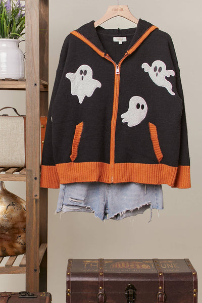 A black knit zip-up hoodie with orange ribbed trim and three white ghost graphics, styled over light blue denim shorts on a wooden hanger against a neutral indoor backdrop.