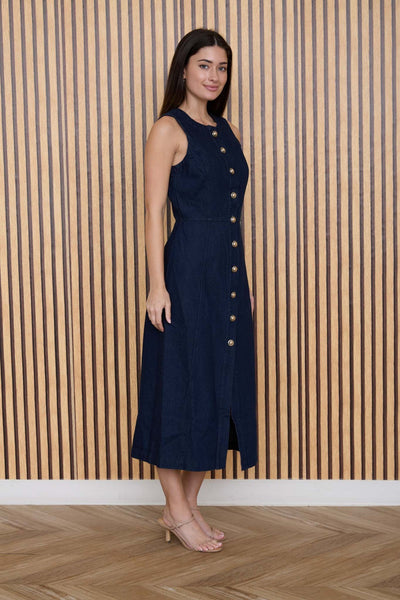 Woman wearing a navy dress with buttons standing against a wooden paneled wall.