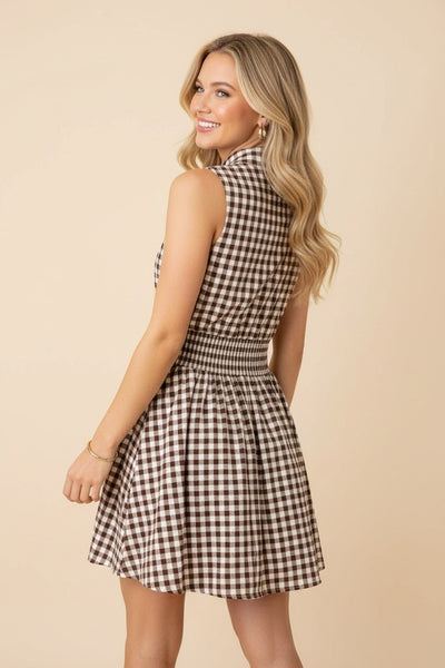 A woman modeling a sleeveless brown and beige gingham mini dress, shown from a three-quarters back angle against a solid beige studio background.