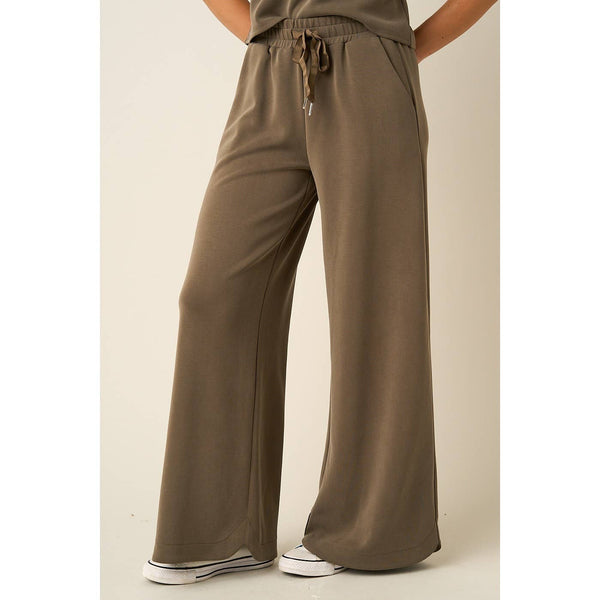A person wearing loose, wide-leg drawstring pants in a muted taupe-brown color, paired with white sneakers, shown from the waist down against a plain beige studio background.