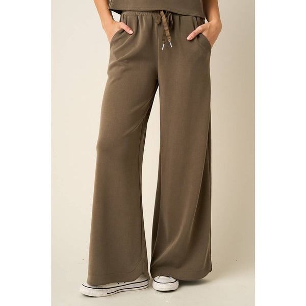 A person wearing wide-leg drawstring pants in a muted taupe-brown color, paired with white sneakers, shown from the waist down against a plain light beige studio background.