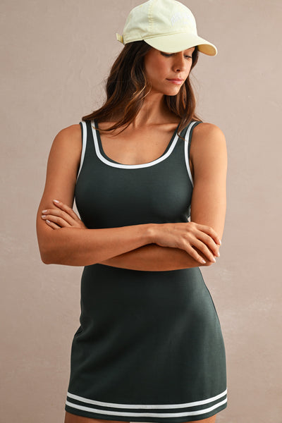 A woman wearing a dark green sleeveless mini dress with white trim and a matching light green baseball cap, standing against a plain beige wall with her arms crossed.