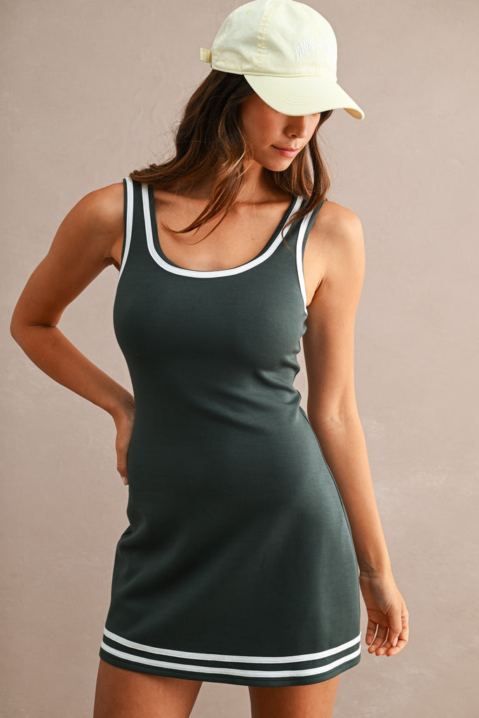 A woman wearing a dark green sleeveless mini dress with white trim and a white baseball cap, posing against a solid beige background.