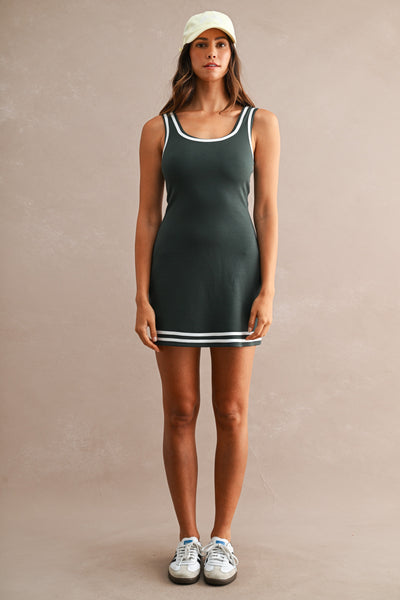 A woman stands against a plain beige studio backdrop wearing a sleeveless dark green mini dress with white trim, a light green cap, and white sneakers.